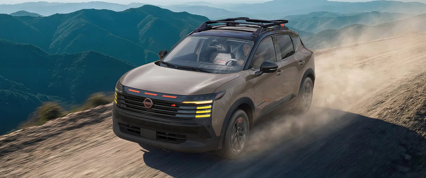 A tan Nissan SUV with a roof rack drives along a dusty dirt road on a mountain ridge.