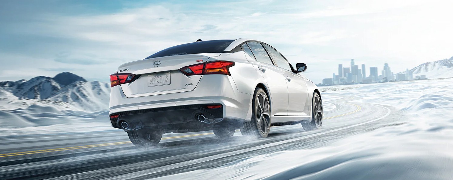 White Nissan Altima SR AWD driving on a winding snowy road with a city skyline in the background.