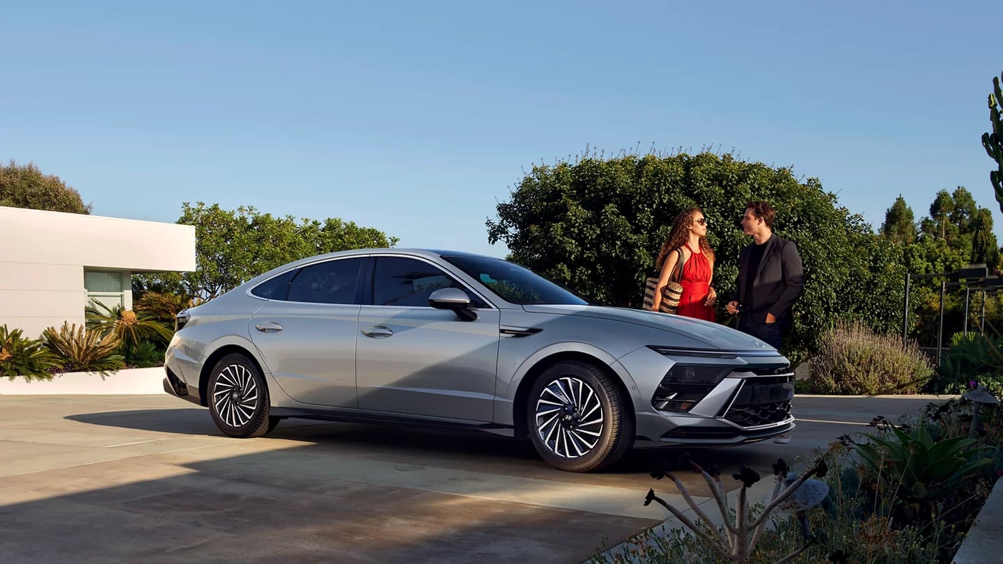 A silver Hyundai Sonata sedan parked on a driveway while a couple stands and chats nearby in daylight.