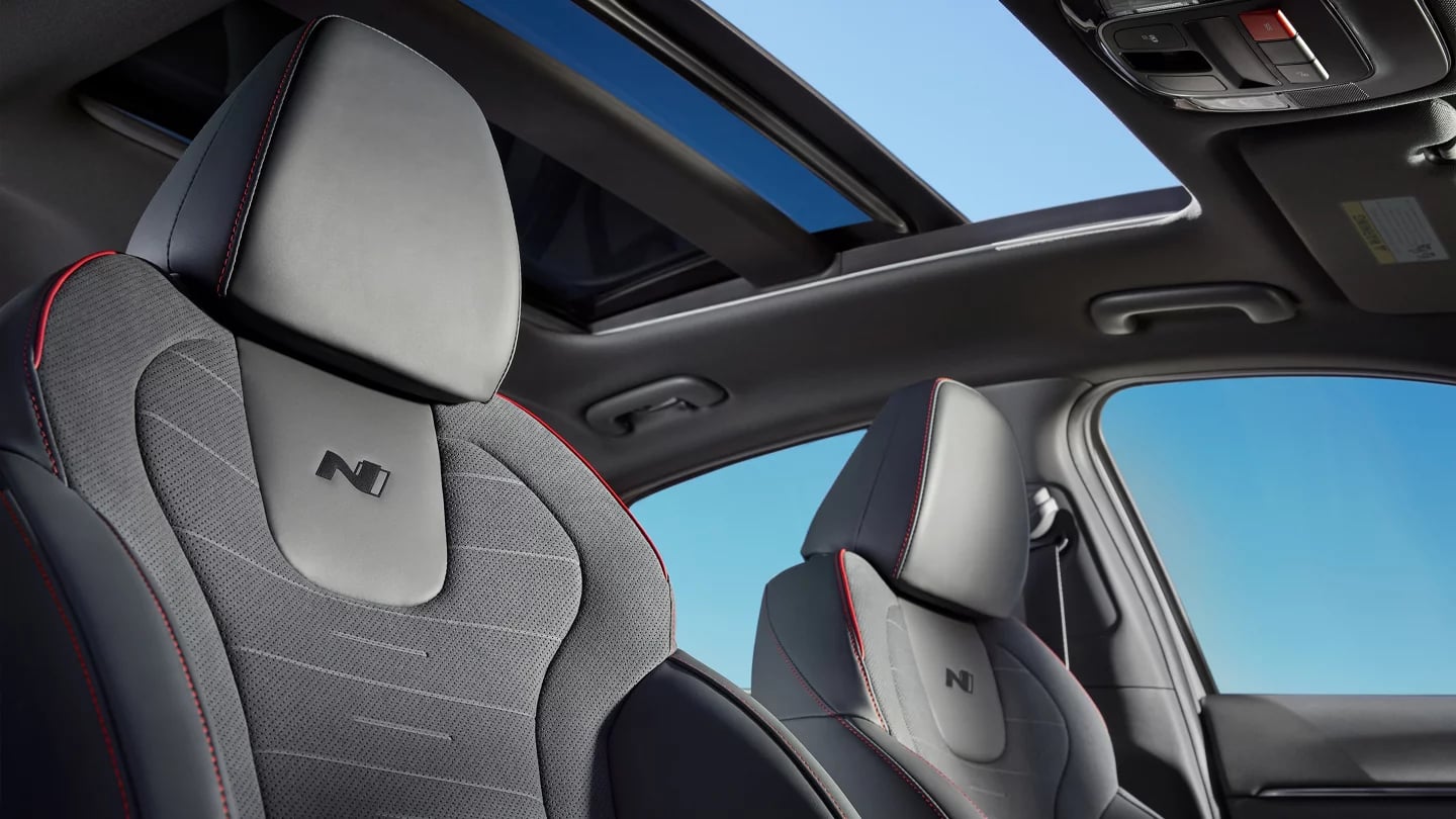 Front sport seats with red stitching and N emblems inside a car under an open panoramic sunroof.