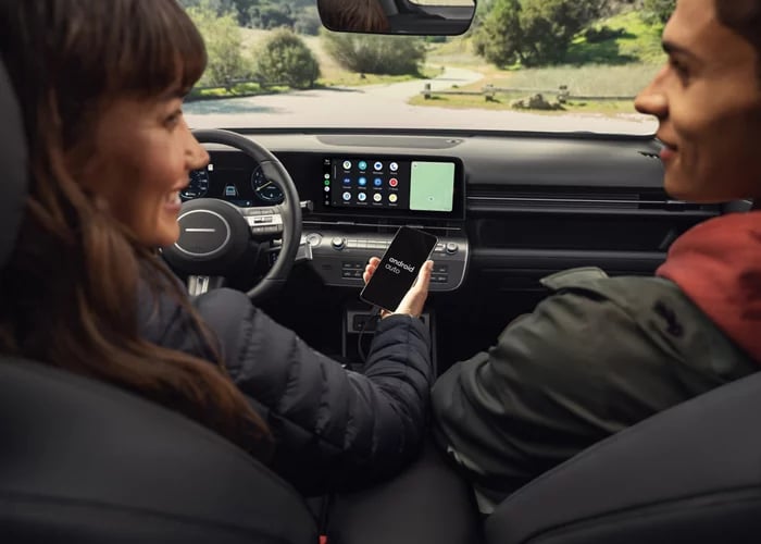 A driver holds a smartphone displaying the Android Auto logo inside a vehicle next to a passenger.