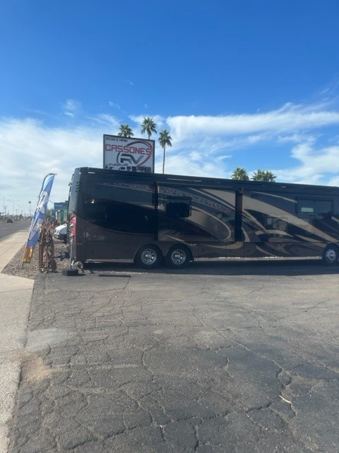 Large black and brown RV parked next to a sidewalk under a blue sky with palm trees.