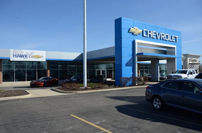 Exterior view of a Chevrolet dealership building with a blue sign tower, banner, and parked cars under a clear sky.