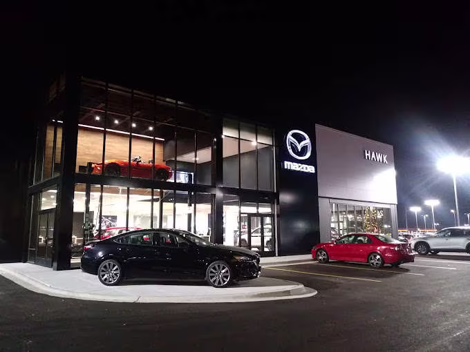 A Hawk Mazda dealership at night with cars on display in the showroom and parking lot.