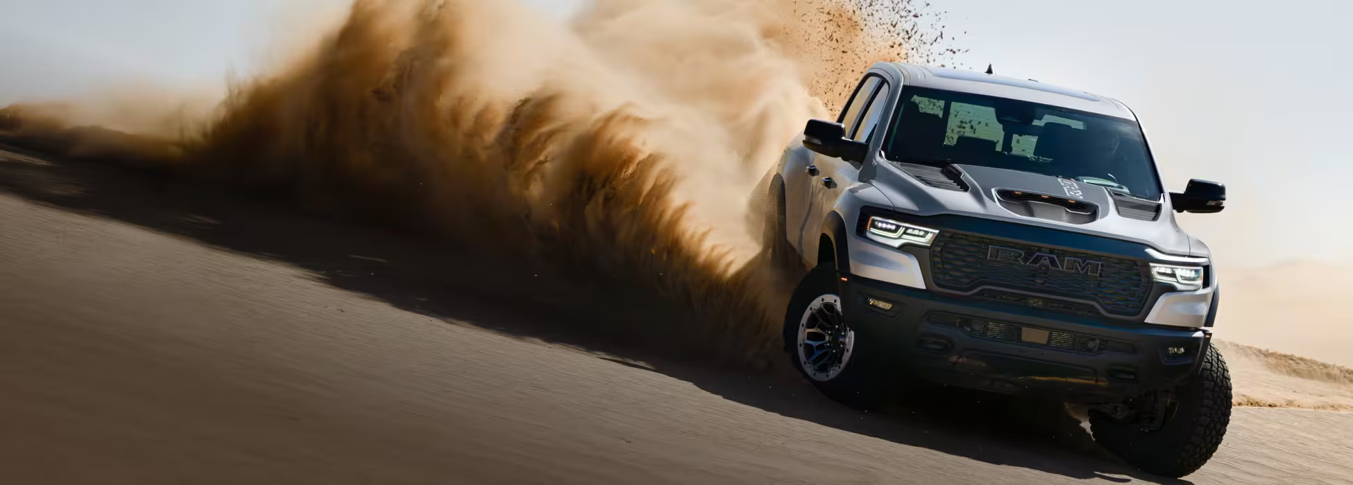 Silver Ram pickup truck driving fast on a desert dune and spraying a cloud of sand into the air.