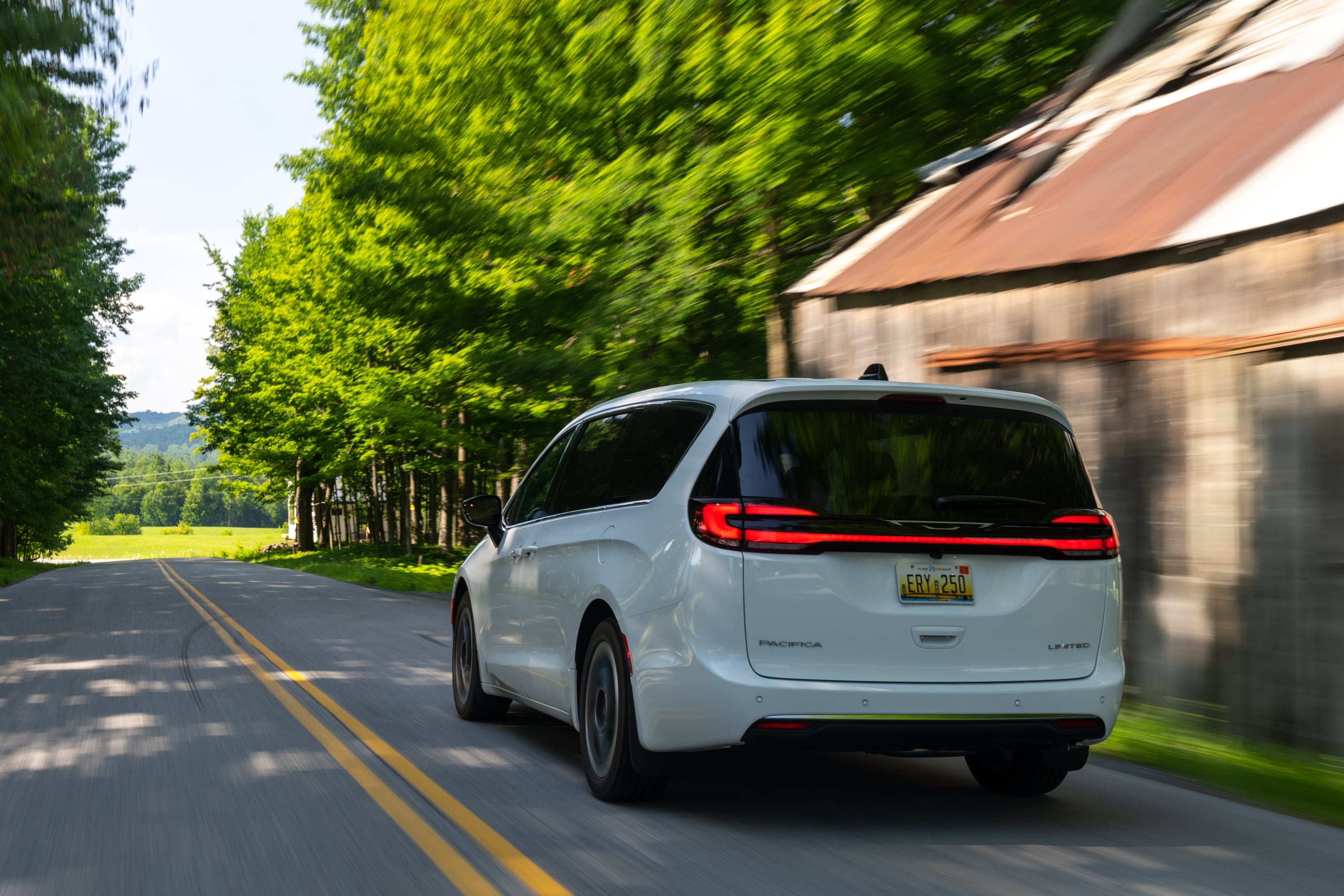 Rear view of a white Chrysler Pacifica minivan driving along a tree-lined rural road past a rustic barn.