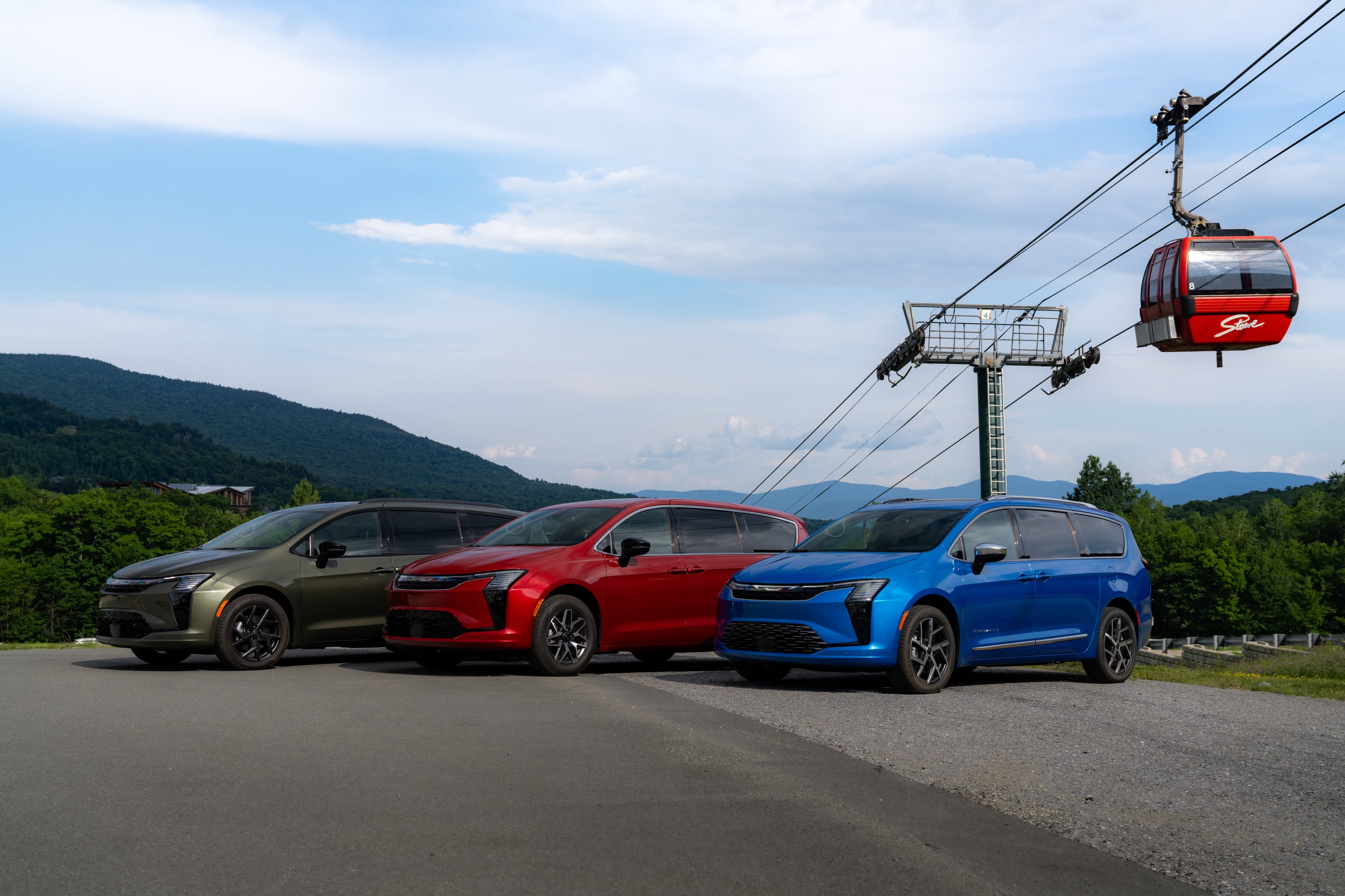 Three minivans in green, red, and blue parked outdoors next to a ski lift gondola with mountains in the background.