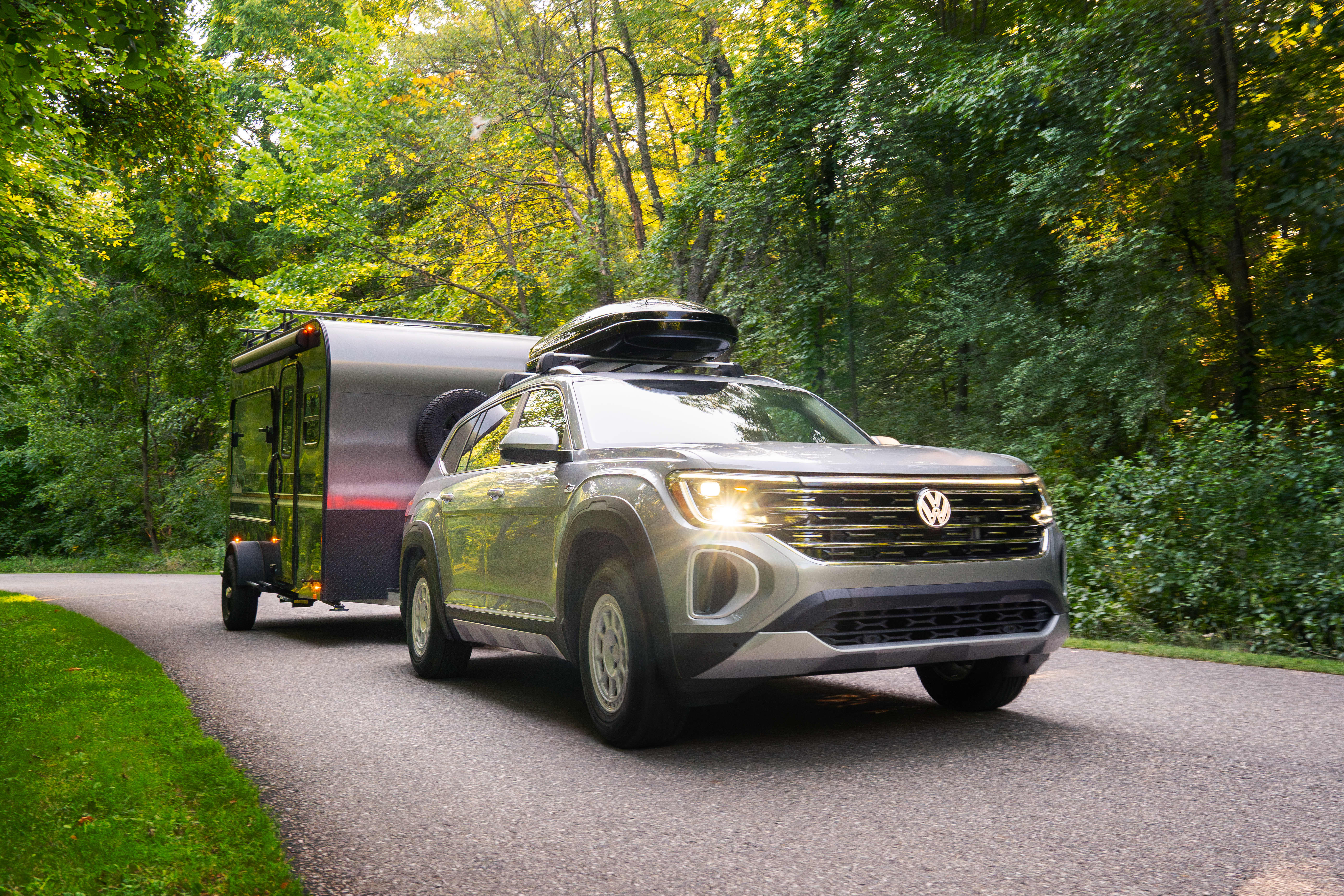 Silver Volkswagen Atlas towing trailer on wooded road, roof cargo carrier.