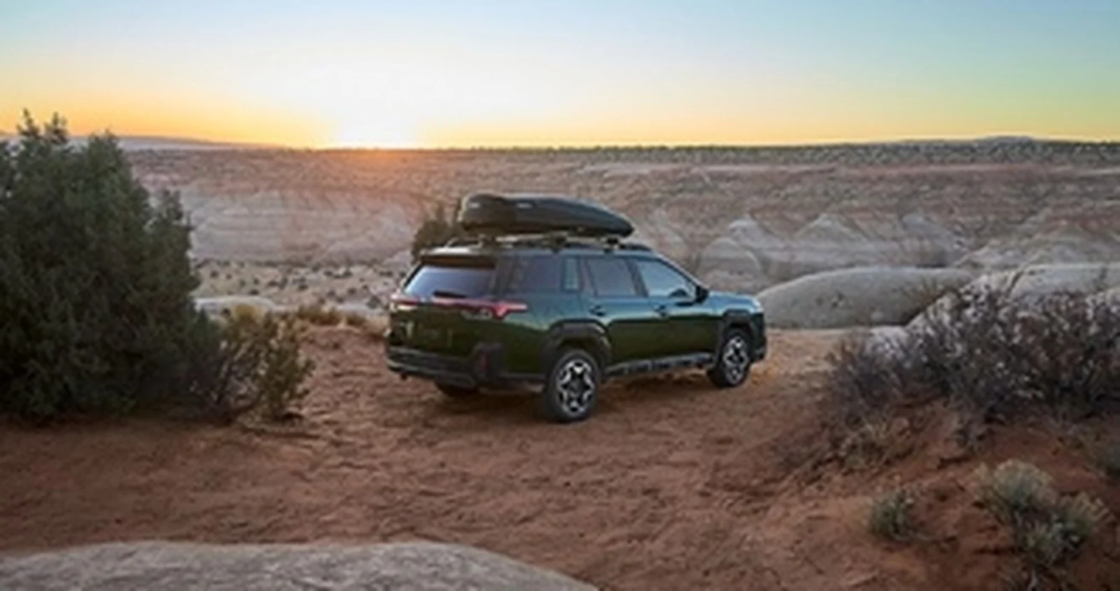 Green Subaru Outback SUV with rooftop cargo carrier in a desert landscape at sunset.