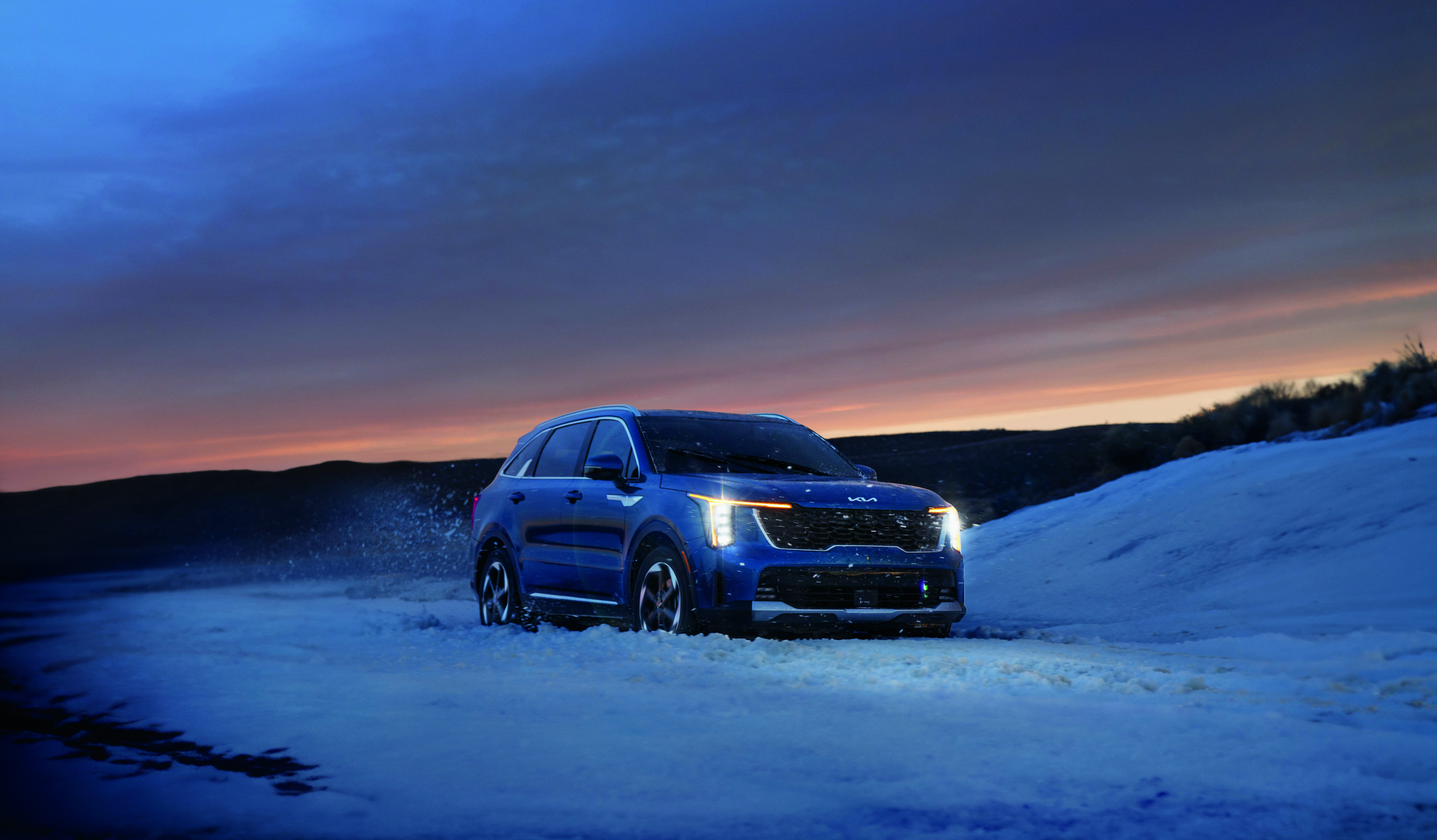 Blue Kia SUV driving through snow at sunset.