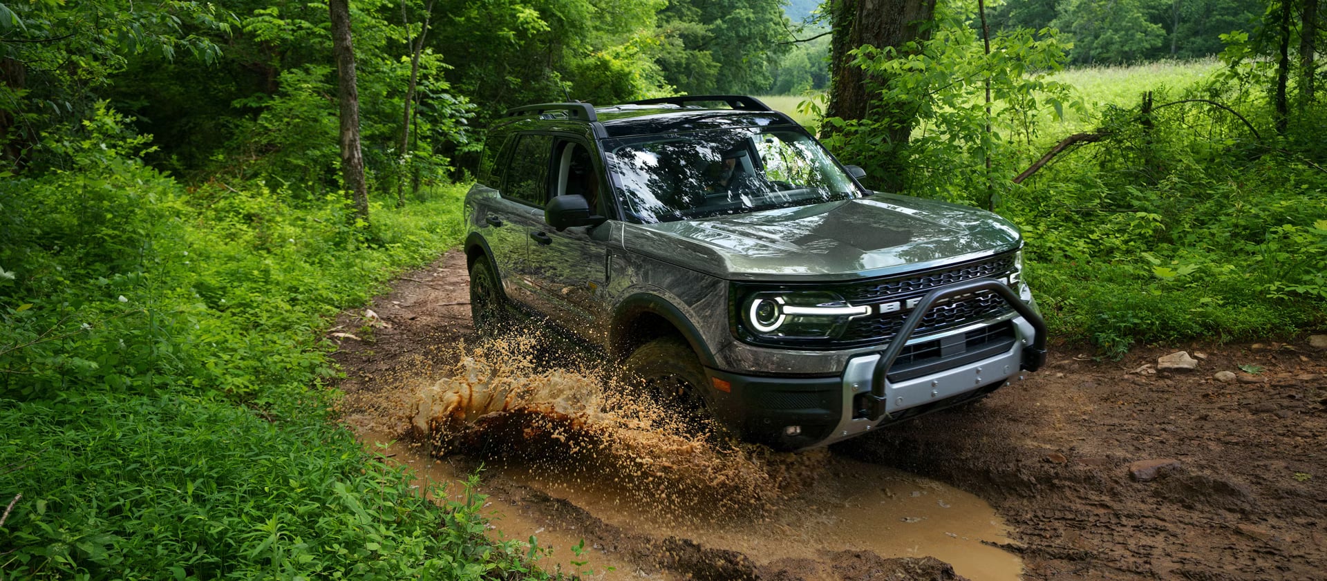 A green Ford Bronco Sport splashing through a muddy puddle on a dirt road in a forest.