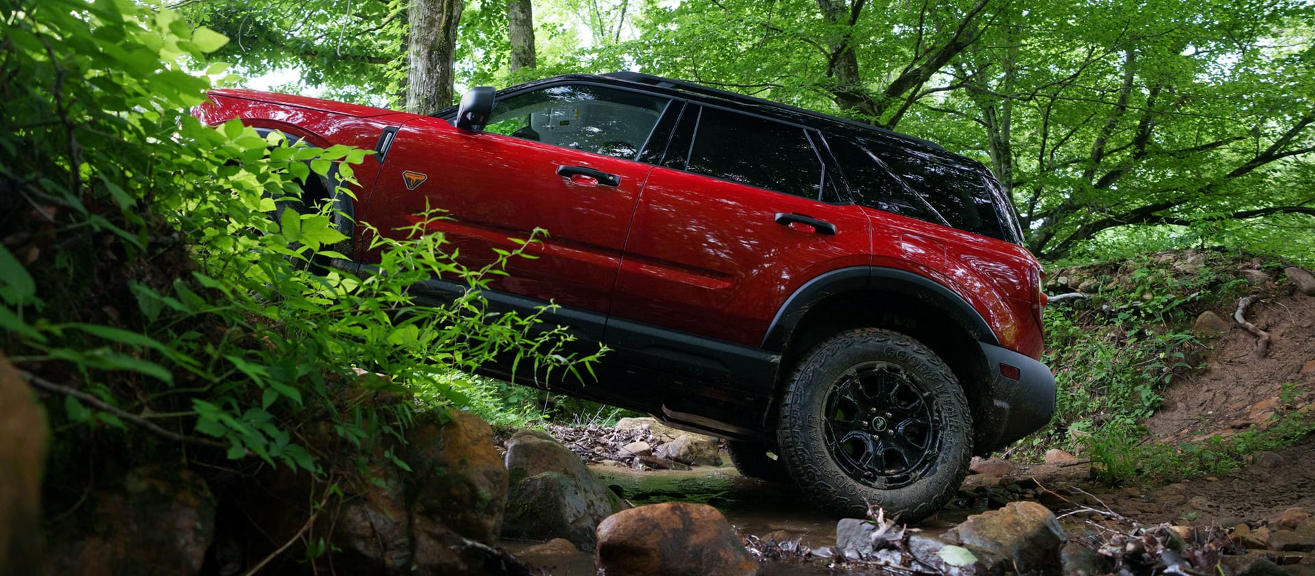 A red Ford Bronco Sport in profile driving over large rocks and a small stream bed.