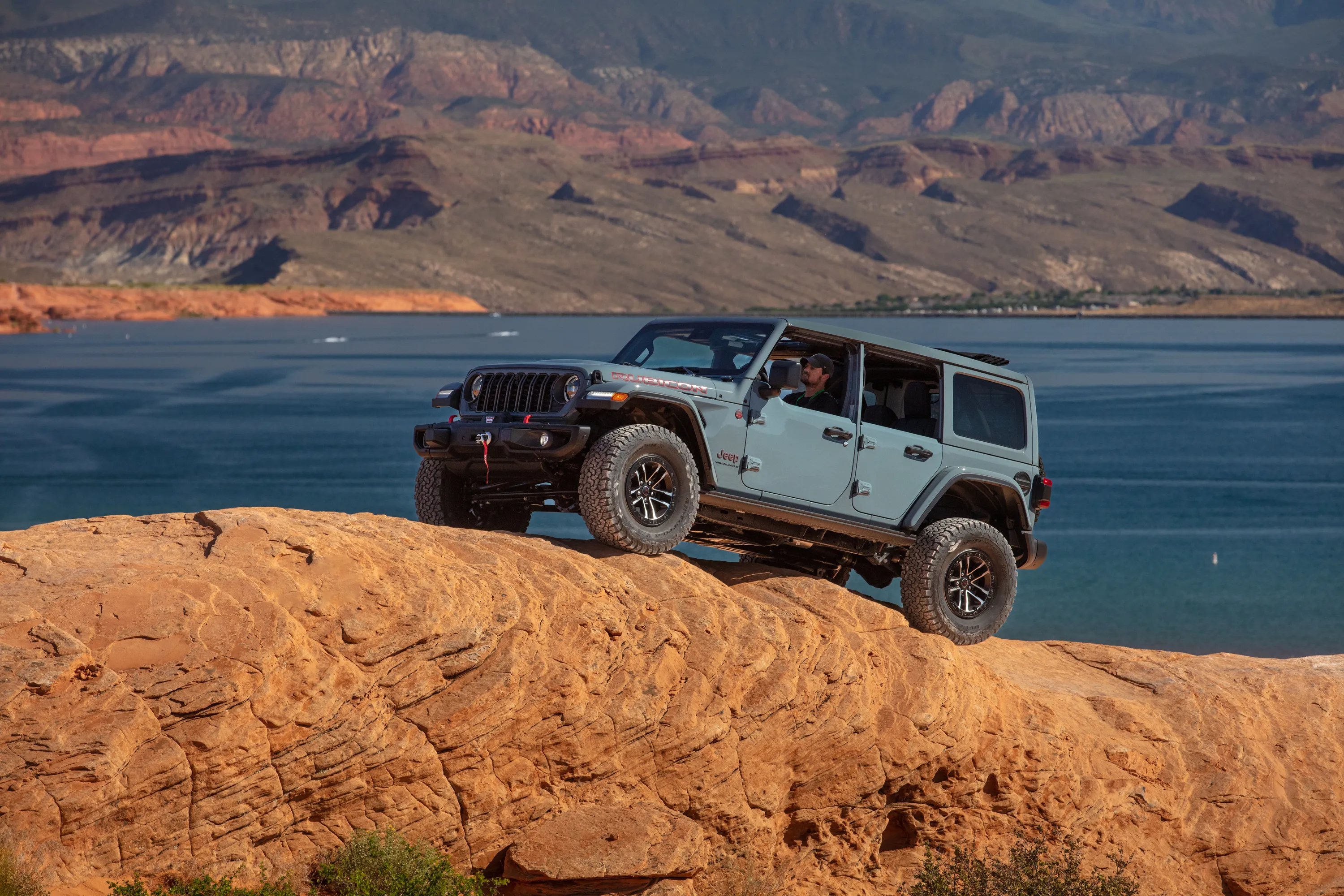 Gray Jeep Rubicon driving on red rock overlooking lake and mountains.