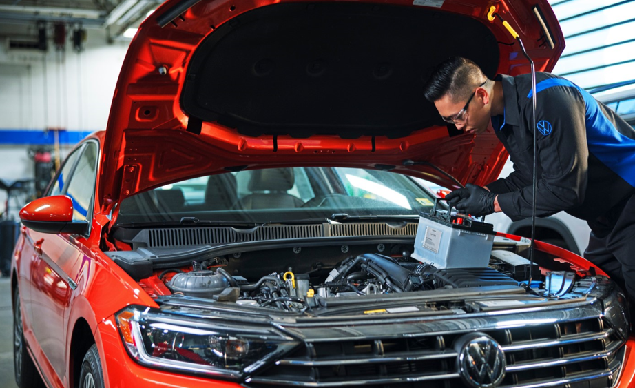 Mechanic replacing a car battery in a red Volkswagen with the hood open.