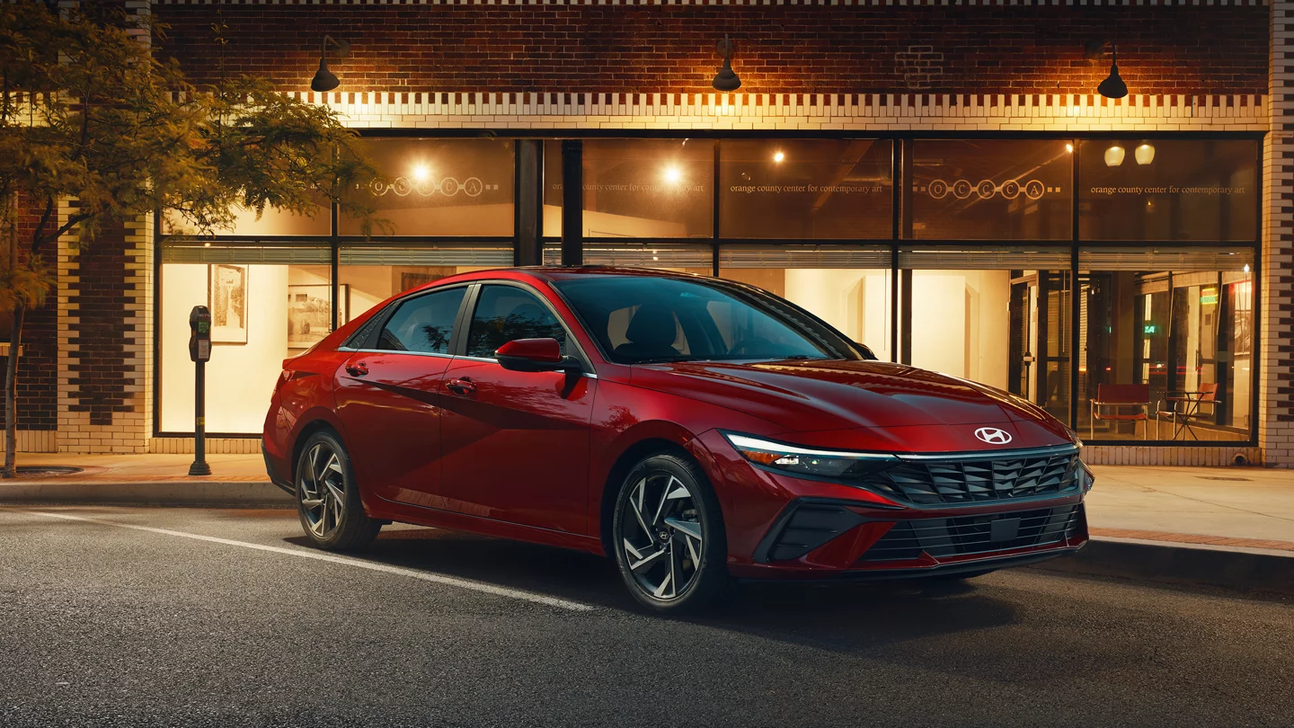 Red Hyundai Elantra parked on a city street at dusk in front of a brick building.