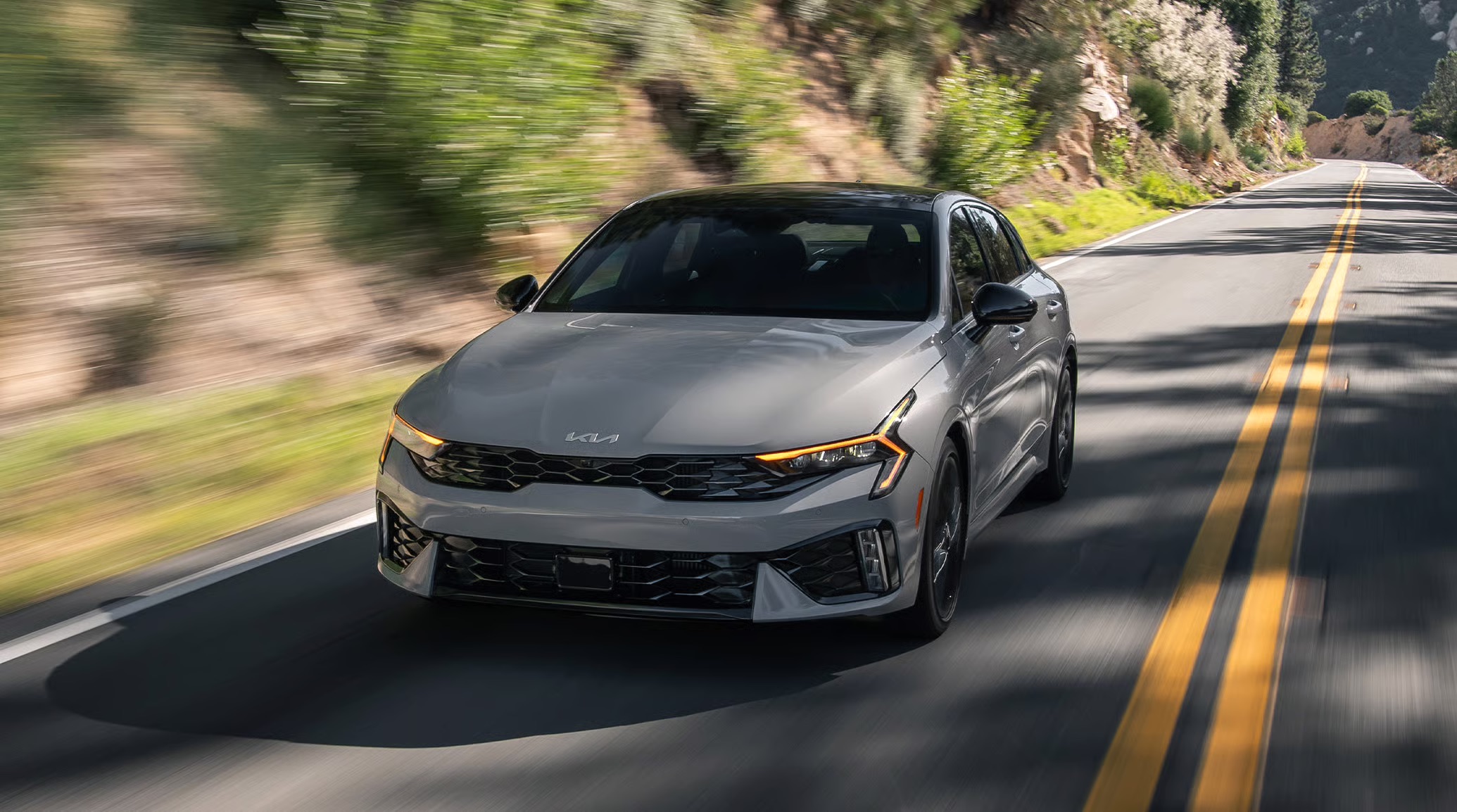 Silver 2026 Kia K5 sedan driving on a winding road.