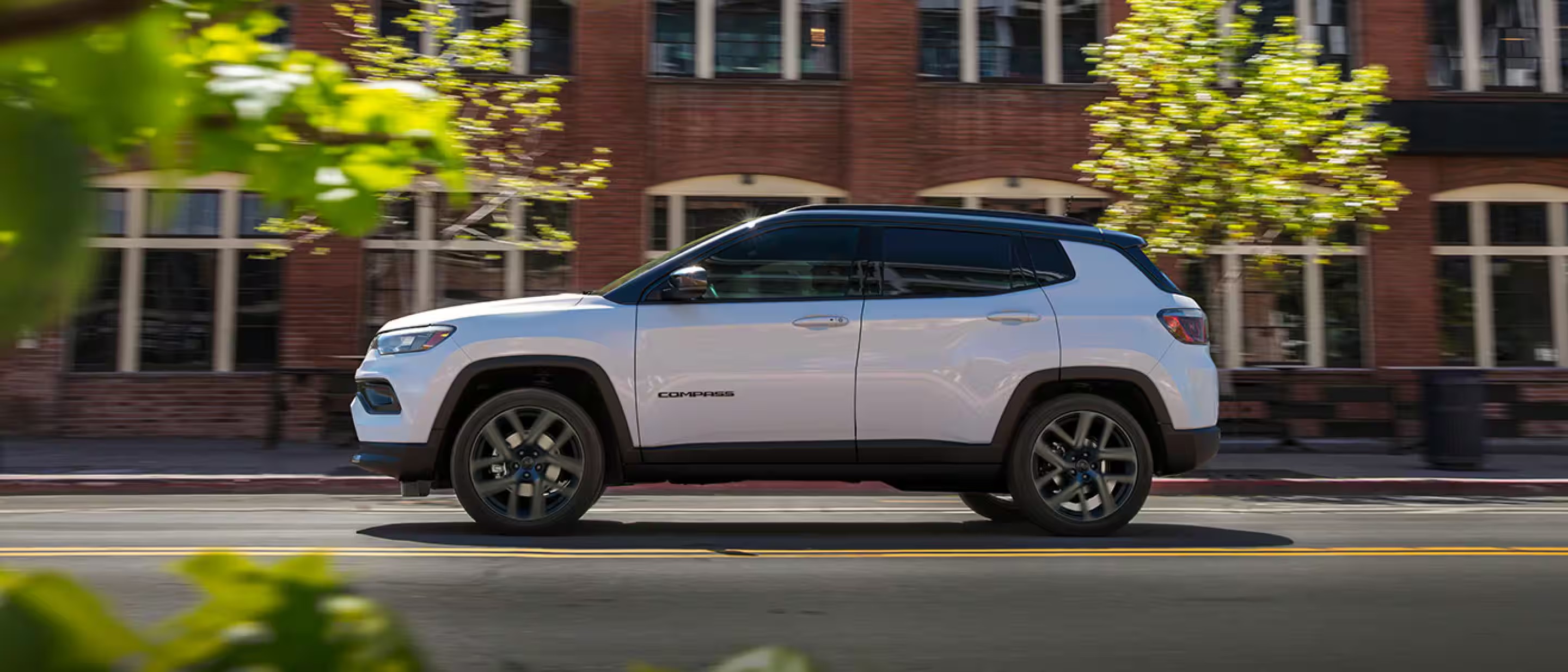White Jeep Compass SUV driving past a brick building on a city street.