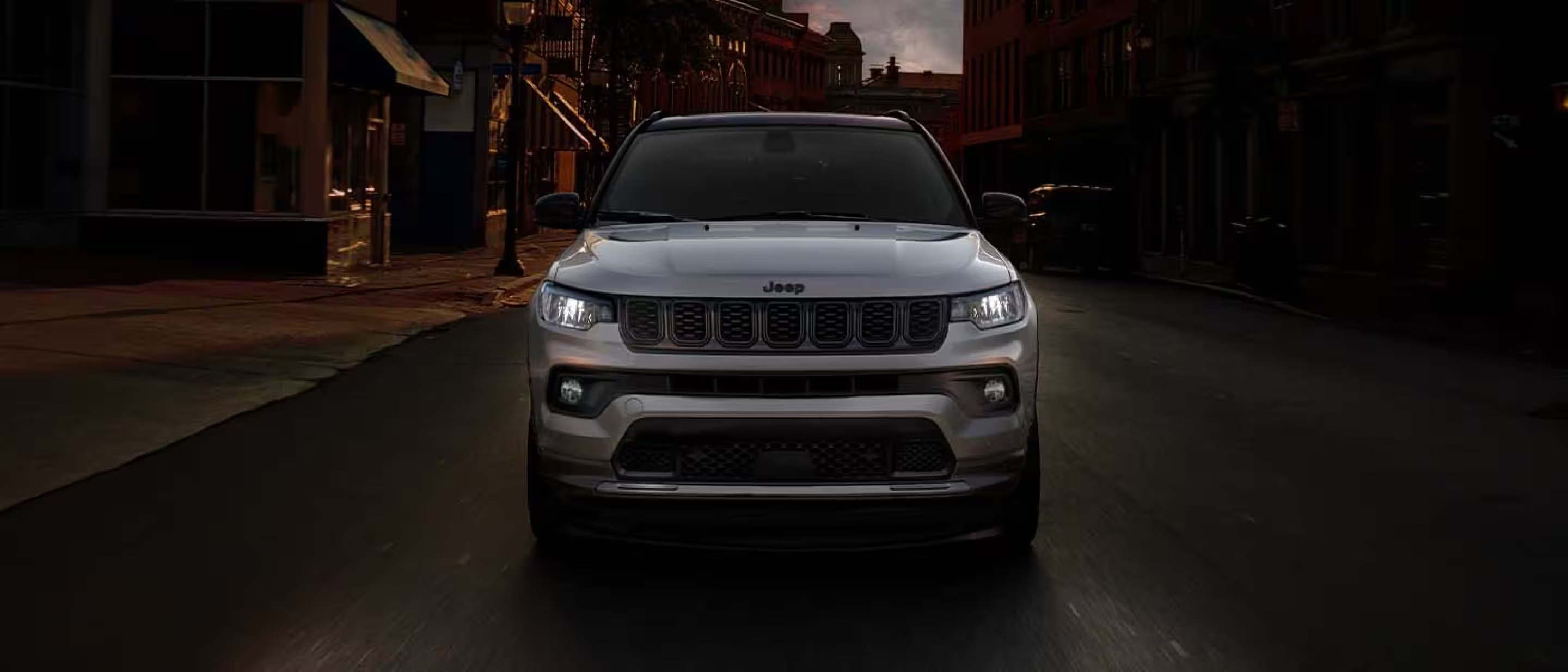 Front view of a silver Jeep Compass driving through a historic city street at sunset.