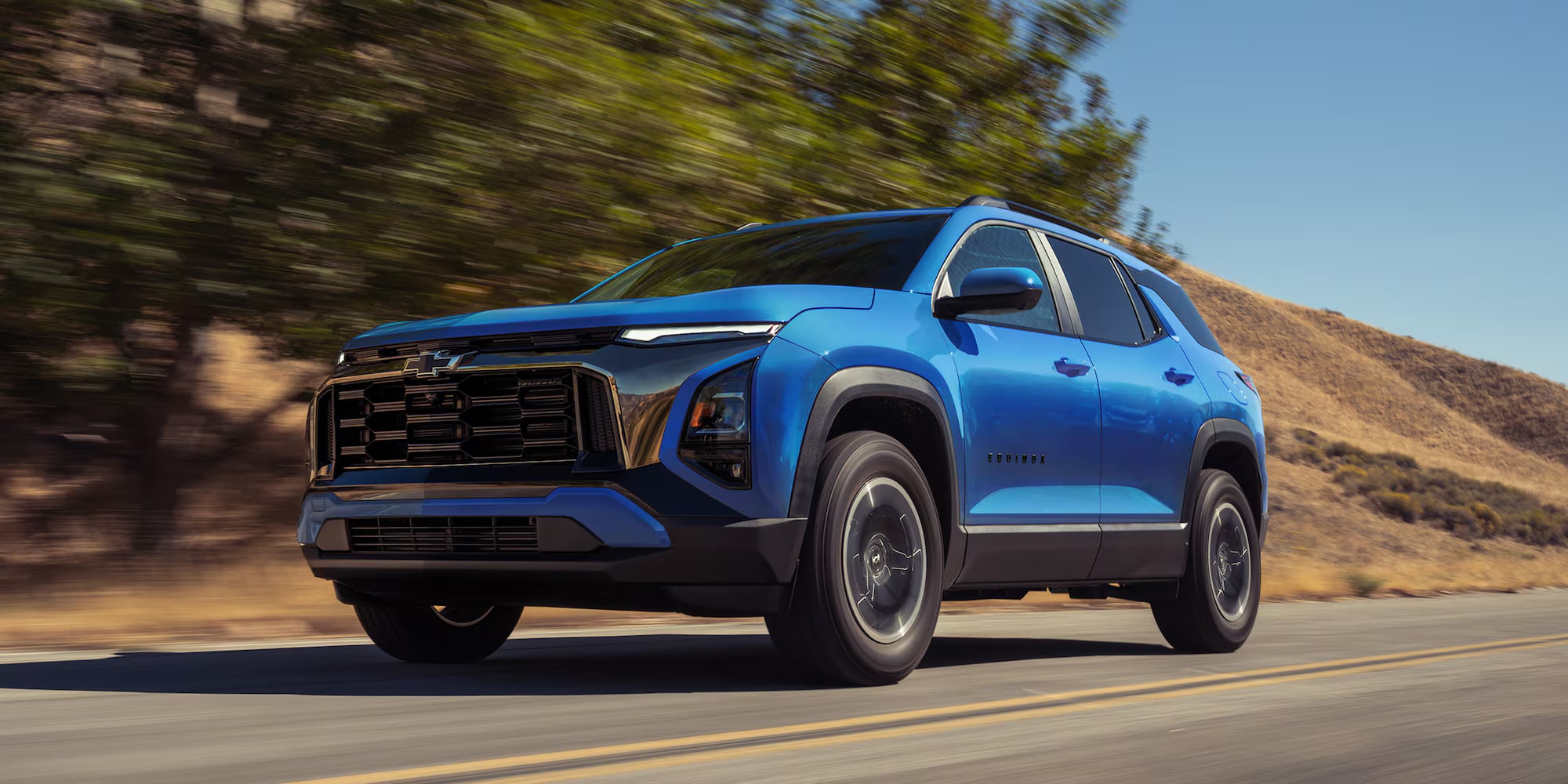 A bright blue Chevrolet Equinox SUV driving along a paved road past sunny hills.