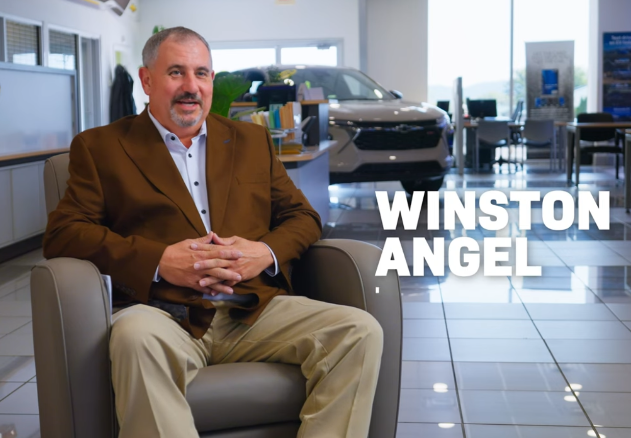 Winston Angel in a brown blazer sitting in a car dealership showroom.