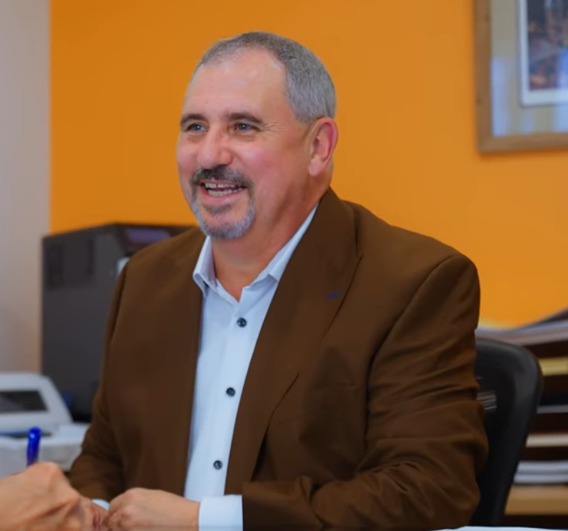 A man with graying hair and a goatee wearing a brown blazer and white shirt smiling at a desk.