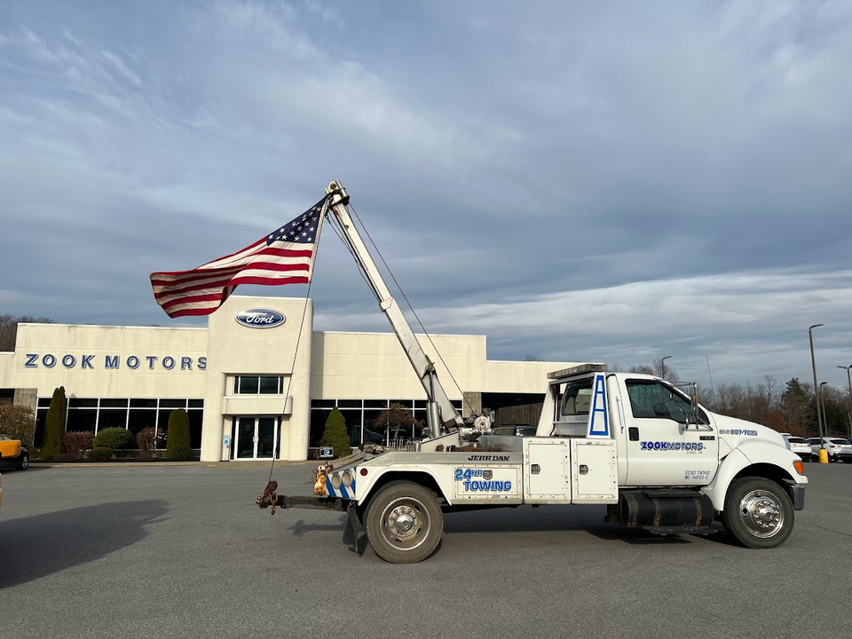A white Zook Motors tow truck with an American flag flying from its extended boom in front of a Ford dealership.