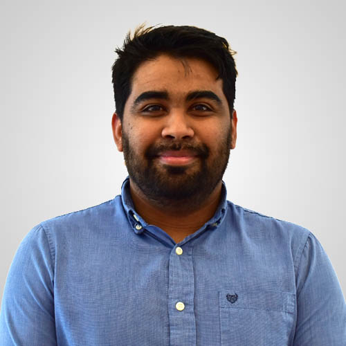 A smiling man with a short beard and dark hair wearing a blue button-down shirt against a grey background.
