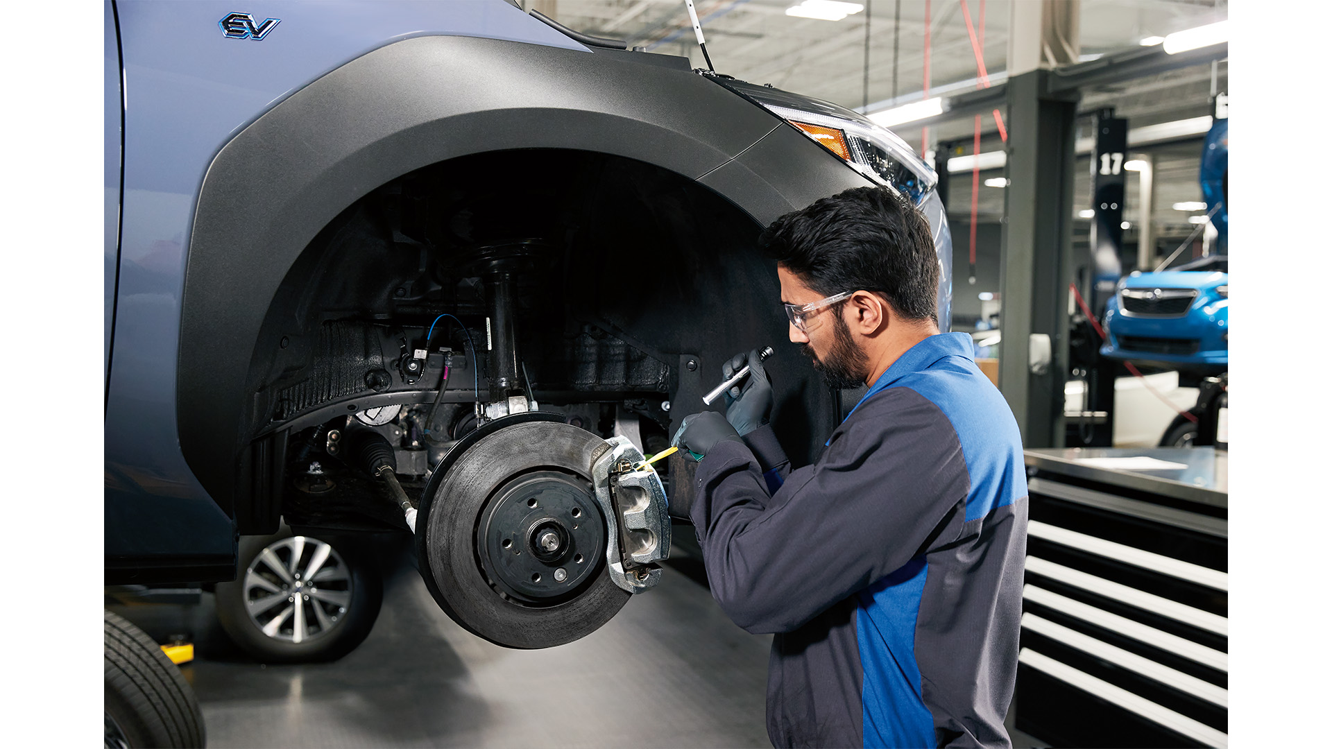 Mechanic in a blue uniform inspecting the brake system of a light blue electric vehicle on a lift.