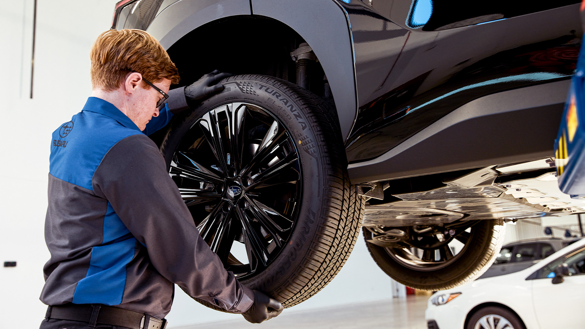 A mechanic installing a black multi-spoke wheel on a vehicle lifted in a service bay.