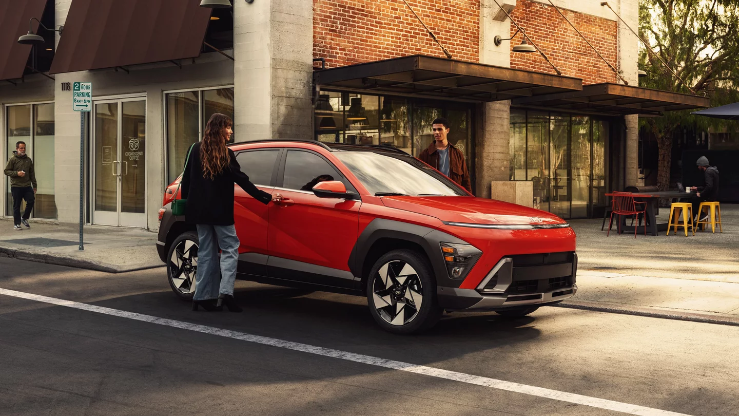 A person opening the door of a red Hyundai Kona on a sunny city street.