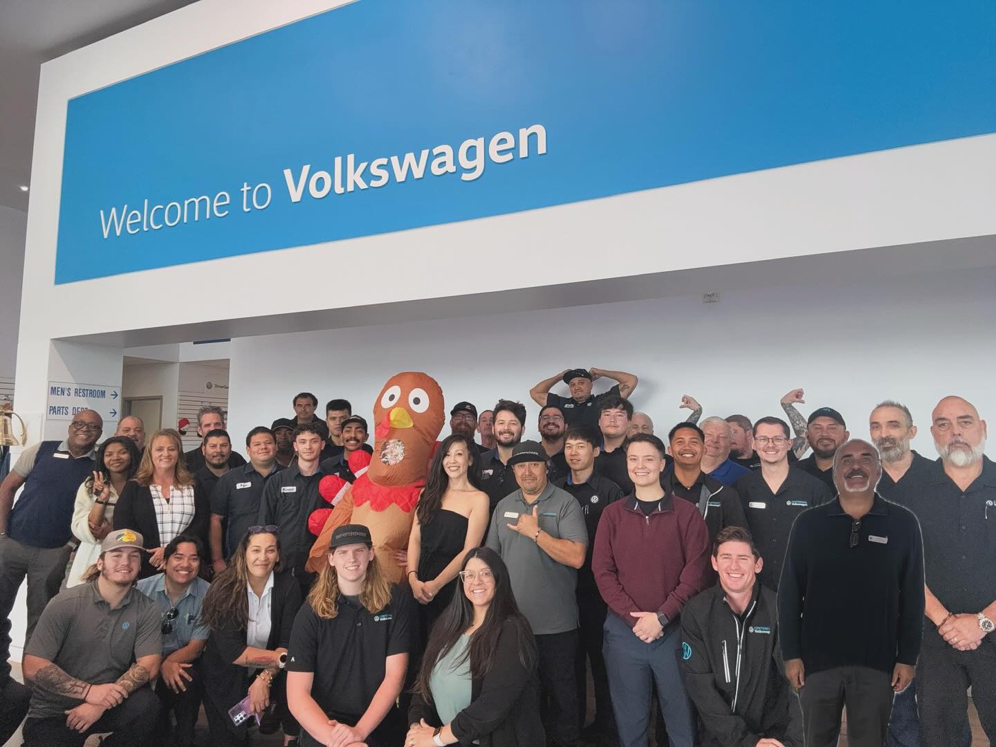 Group photo of Volkswagen dealership staff, including a person in a turkey costume, posing under dealership sign.