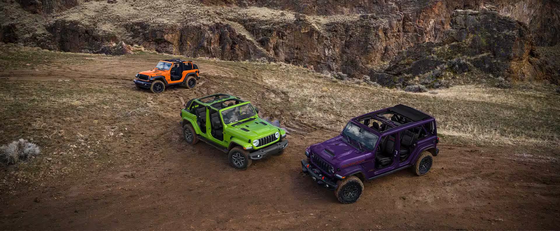 Three open-top Jeep Wranglers in orange, green, and purple parked on a dirt path in a rocky canyon.