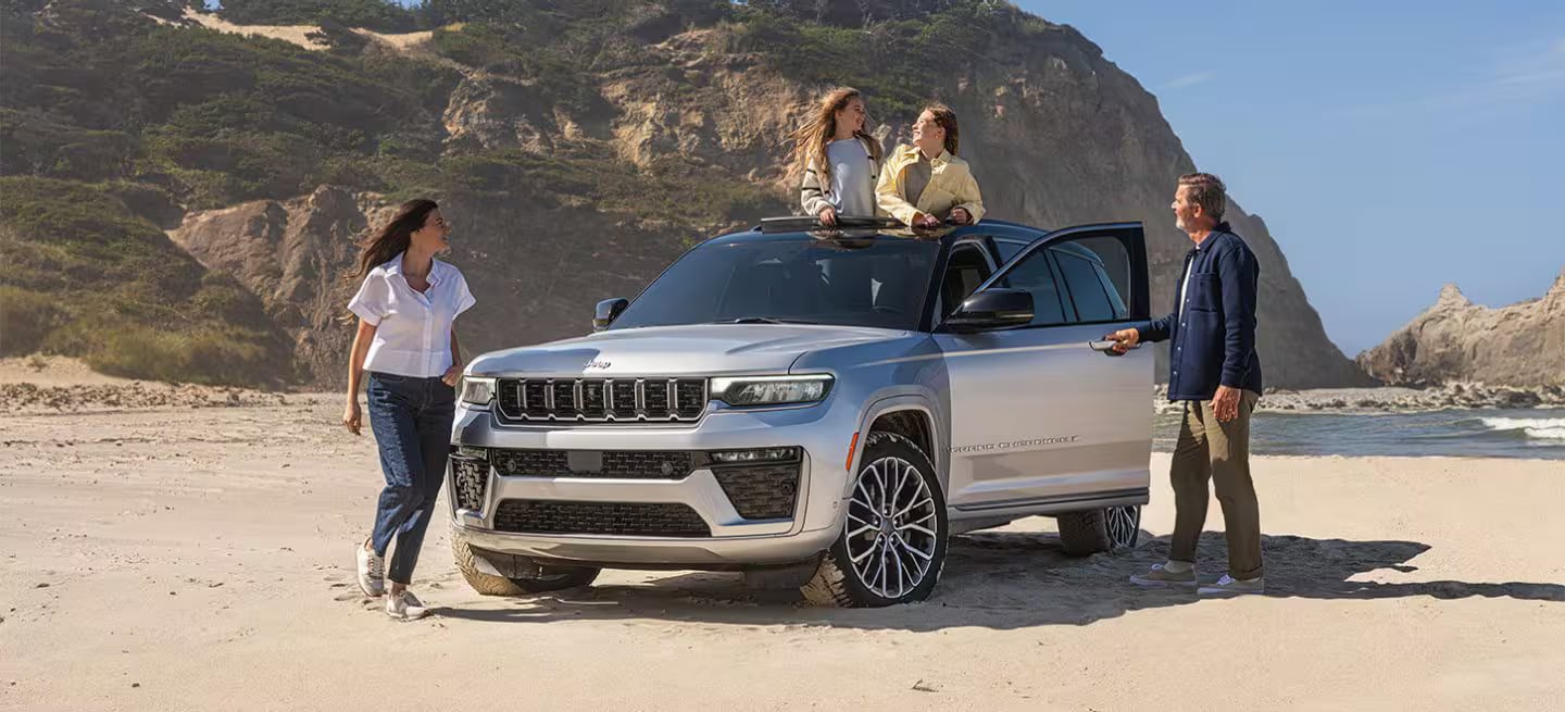 A family of four enjoying a beach day with their silver Jeep Grand Cherokee on a sandy shore.