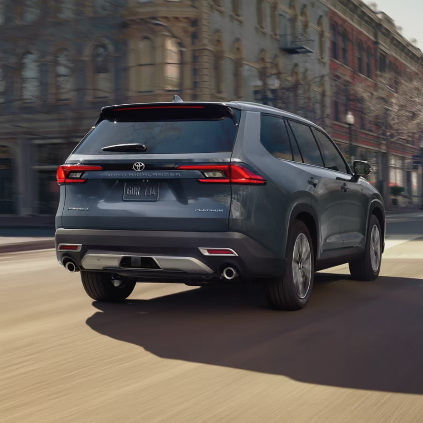 Gray Toyota Grand Highlander driving down a city street from the rear three-quarter angle.