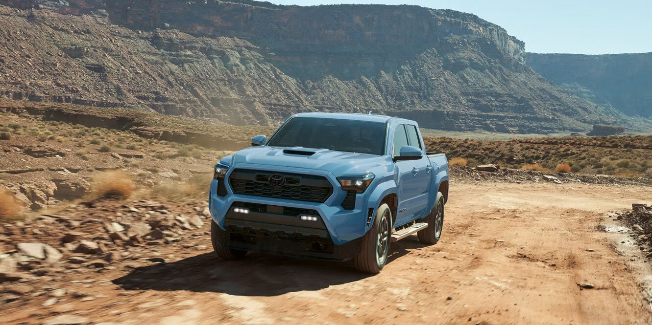 A light blue Toyota Tacoma pickup truck driving along a dirt road through a desert canyon landscape.