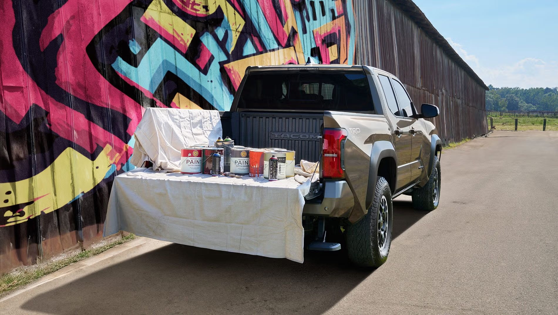 Bronze Toyota Tacoma parked beside a graffiti wall with paint cans arranged on its open tailgate.