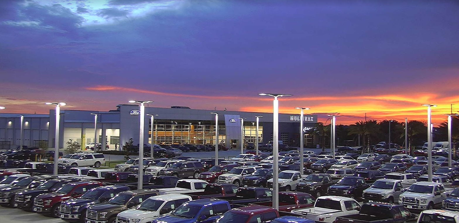 A Ford car dealership lot filled with pickup trucks under a vibrant sunset sky.