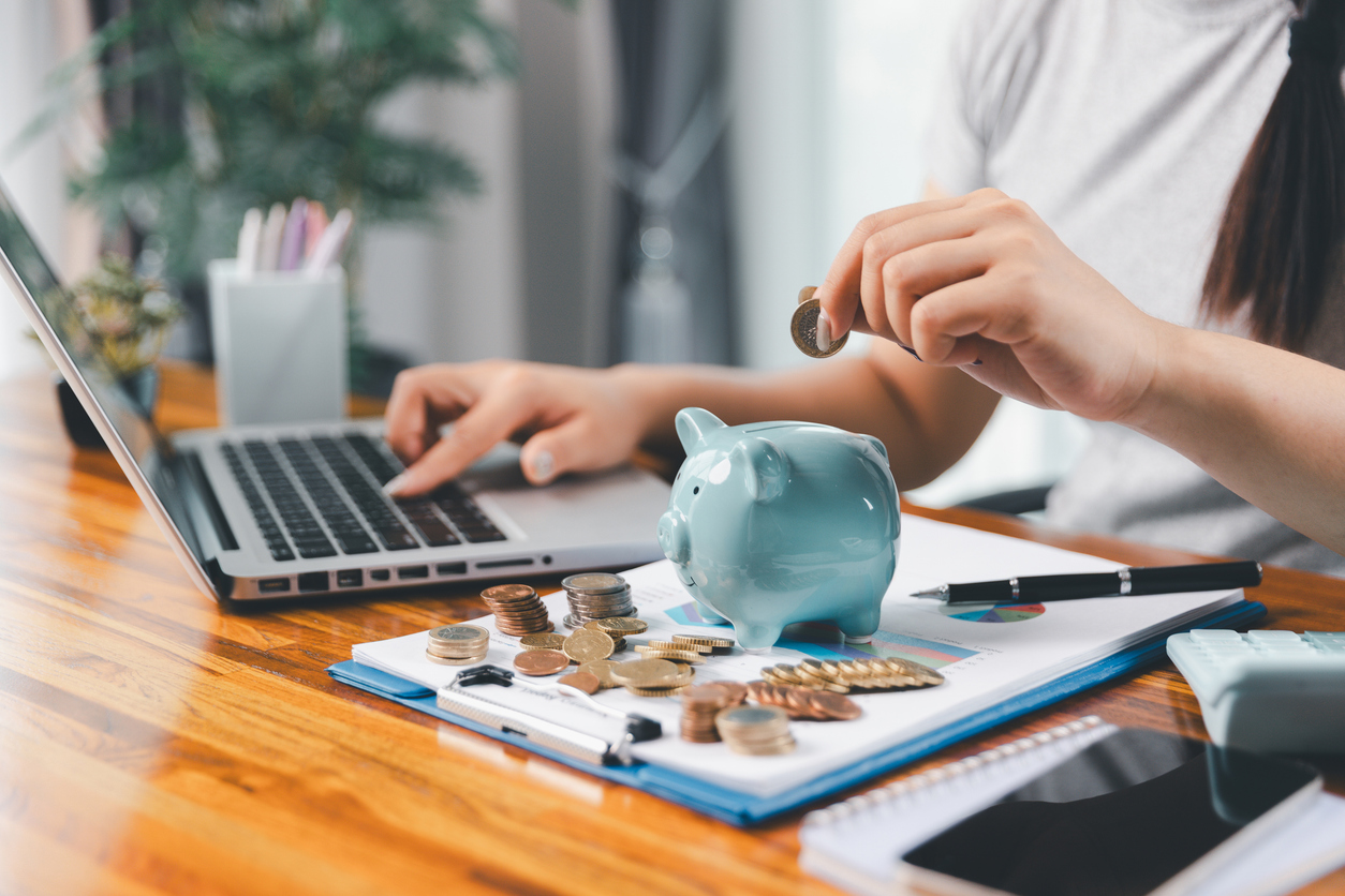 A person dropping a coin into a blue piggy bank while using a laptop at a desk with money and financial charts.
