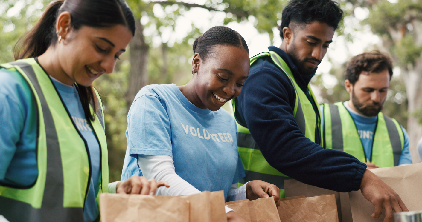 Four diverse volunteers in blue shirts and neon vests pack brown paper bags outdoors.