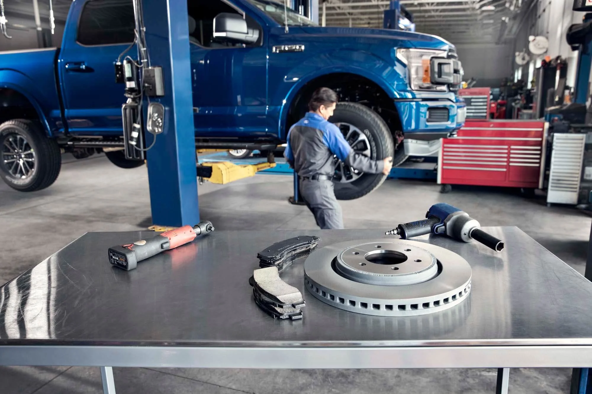 A mechanic working on a blue truck in a garage with brake parts on a foreground table.